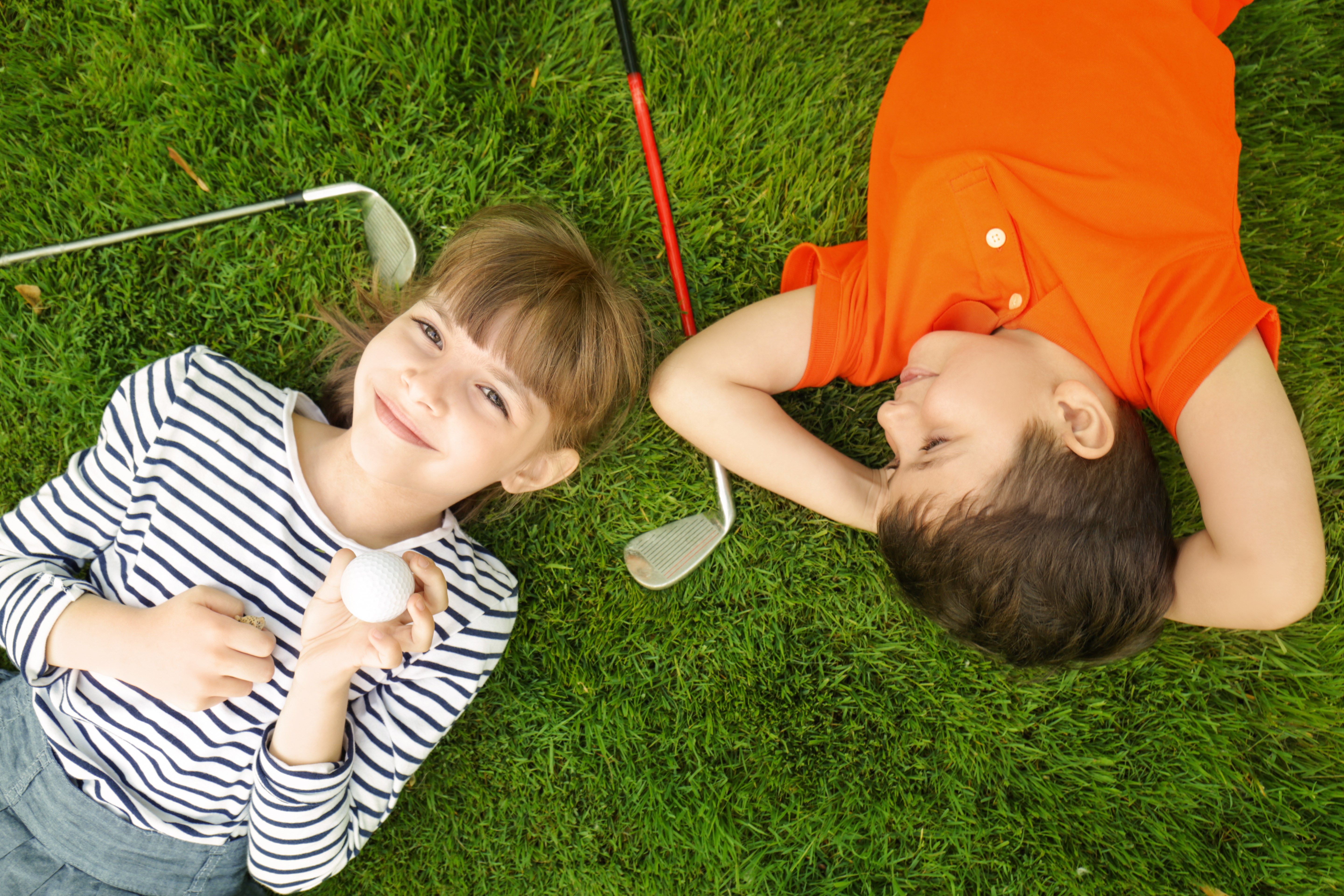 Två barn ligger på en golfbana och har två golfklubbor bredvid sig.